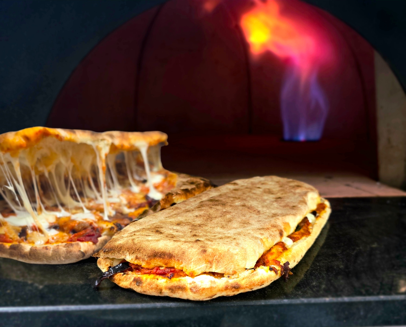 Cheese-pull pizza slice and folded panuozzo in front of the glowing wood-fired oven